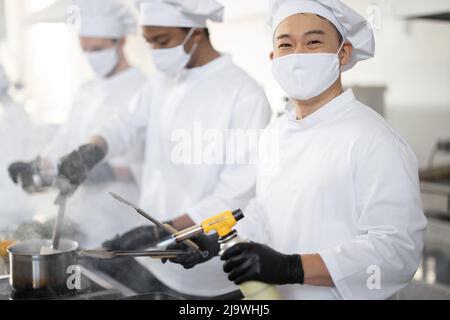 Multirassisches Team von Köchen kocht in weißer Uniform zusammen in der Küche. Asiatischer Koch, der Fleisch mit Brenner, lateinamerikanische und europäische Küche auf dem Hintergrund braten lässt. Köche tragen Gesichtsmasken und Schutzhandschuhe Stockfoto