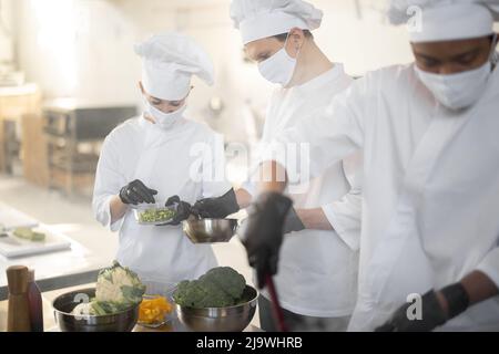 Multirassisches Team von Köchen kocht in weißer Uniform zusammen in der Küche. Asiatische, lateinische und europäische Jungs kochen zusammen. Köche tragen Gesichtsmasken und Schutzhandschuhe Stockfoto