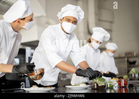 Mehrere Rassen Gruppe von Köchen, die Hauptgerichte beenden, während sie zusammen in der Küche arbeiten. Köche tragen Uniform und Gesichtsmaske. Das Team bereitet während einer Pandemie Mahlzeiten für das Restaurant zu Stockfoto