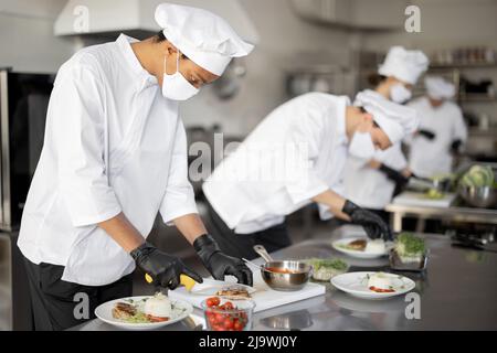 Mehrere Rassen Gruppe von Köchen, die Hauptgerichte beenden, während sie zusammen in der Küche arbeiten. Köche tragen Uniform und Gesichtsmaske. Das Team bereitet Mahlzeiten für das Restaurant zu Stockfoto