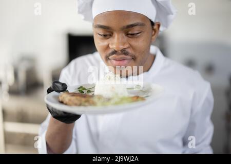 Porträt eines jungen, gutaussehenden, lächelnden Küchenchefs in Uniform, der mit einer Fertigmahlzeit in der Küche steht. Fröhlicher lateinamerikanischer Koch, der im Restaurant arbeitet. Konzept der beruflichen Tätigkeit und Haute Cuisine Stockfoto