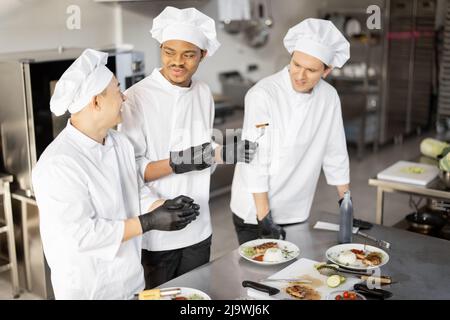 Drei Köche mit unterschiedlichen Nationalitäten verkosten gekochte Gerichte und erfinden neue Gerichte für ein Menü. Asiatischer Koch mit lateinischen und europäischen Köchen in Uniform, die zusammenarbeiten. Konzept der Teamarbeit und hohe Küche Stockfoto
