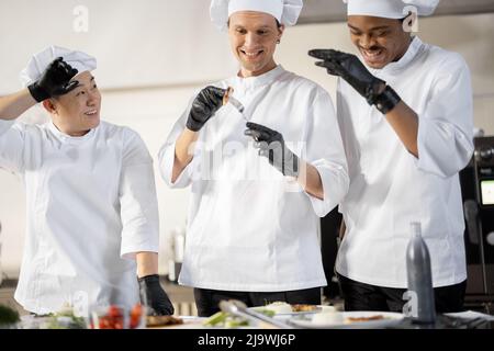 Drei Köche mit unterschiedlichen Nationalitäten verkosten gekochte Gerichte und erfinden neue Gerichte für ein Menü. Kaukasischer Koch mit lateinischen und asiatischen Köchen in Uniform, die zusammenarbeiten. Konzept der Teamarbeit und hohe Küche Stockfoto
