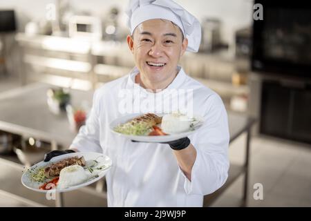 Porträt eines jungen glücklichen, gutaussehenden, lächelnden Küchenchefs in Uniform, der mit einer Fertigmahlzeit in der Küche steht. Glücklicher asiatischer Koch, der im Restaurant arbeitet. Konzept der beruflichen Tätigkeit und Haute Cuisine Stockfoto