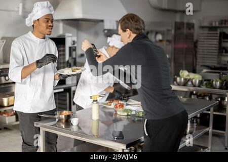 Mann zeichnet am Telefon den Prozess des Kochens von zwei Köchen in einer professionellen Küche auf. Lateinamerikanische und asiatische Köche bereiten Speisen für soziale Medien zu. Konzept des kulinarischen Videobloggens Stockfoto