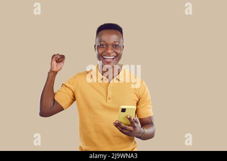 Glücklicher afroamerikanischer Kerl, der sich aufgeregt fühlt, während er Inhalte auf dem Bildschirm seines Mobiltelefons anguckt. Stockfoto
