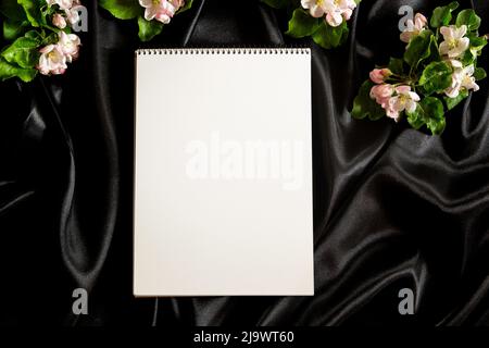 Leere offene Spirale Skizzenbuch Notizbuch und Frühling blühenden Baum Äste mit weißen Blumen auf eleganten schwarzen Seide Satin Stoff Hintergrund. Draufsicht Stockfoto