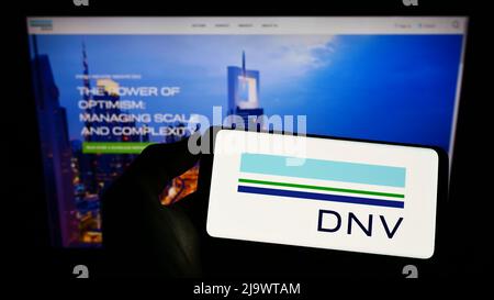 Person, die Mobiltelefon mit dem Logo des norwegischen Dienstleistungsunternehmens DNV AUF dem Bildschirm vor der Business-Webseite hält. Konzentrieren Sie sich auf die Telefonanzeige. Stockfoto