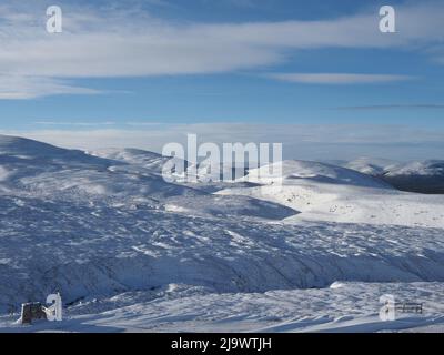 Der Blick auf schneebedeckte Pisten und Berge vom Cairngorm Skigebiet in Richtung Chalamain Gap, mit einem wolkenblauen Himmel und Sonnenschein. Stockfoto