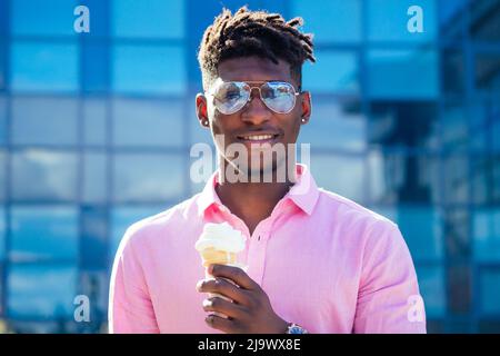 Der coole und modische afroamerikanische Kerl Dreadlocks Haare in Sonnenbrillen und isst im Sommer im Park Eis in einem Waffelhorn Stockfoto