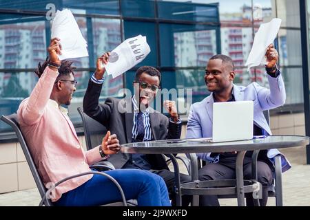Eine Gruppe ihrer drei erfolgreichen afroamerikanischen Geschäftsleute in einem stilvollen Anzug sitzt am Tisch und arbeitet mit einem Laptop und einem Fenster mit Papierhochhäusern Stockfoto