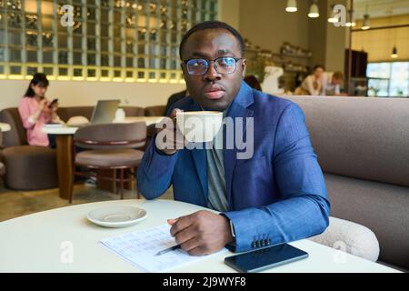 Afrikanischer junger Geschäftsmann im Anzug, der Kaffee trinkt, während er mit Dokumenten und Handy am Tisch im Café sitzt Stockfoto