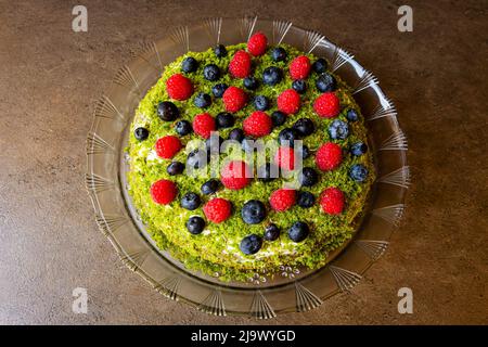 Draufsicht auf einen runden hausgemachten Obstkuchen mit roten Himbeeren, Blaubeeren und grüner Spitze aus Spinat auf Glasschale auf brauner Oberfläche gelegt. Stockfoto