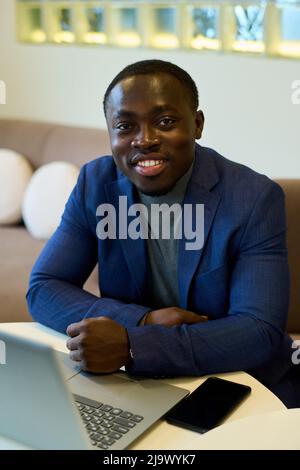 Porträt eines afrikanischen Geschäftsmannes in blauem Anzug, der mit einem Lächeln auf die Kamera lächelt, während er mit einem Laptop am Tisch im Restaurant sitzt Stockfoto