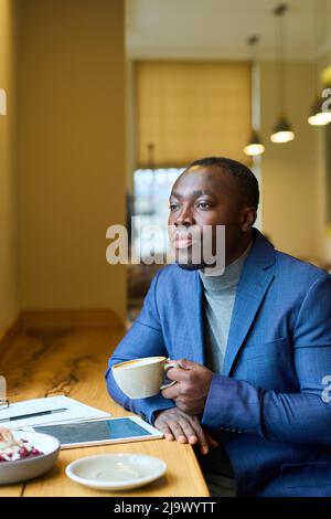 Afrikanischer seriöser Geschäftsmann, der mit einem Tablet-pc am Tisch sitzt und nachdenklich aussieht, während er im Café Kaffee trinkt Stockfoto