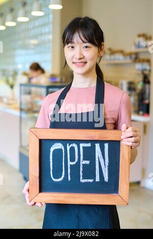 Porträt der jungen asiatischen Kellnerin in Uniform halten Schild und lächeln an der Kamera, sie Eröffnung Café Stockfoto