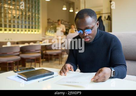 Afrikanischer junger Geschäftsmann in Brillen, der das Menü am Tisch im Café untersucht Stockfoto