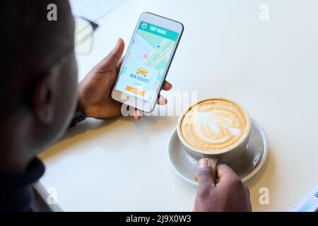 Nahaufnahme eines Mannes, der am Tisch Kaffee trinkt und mit seinem Mobiltelefon ein Taxi bestellt Stockfoto