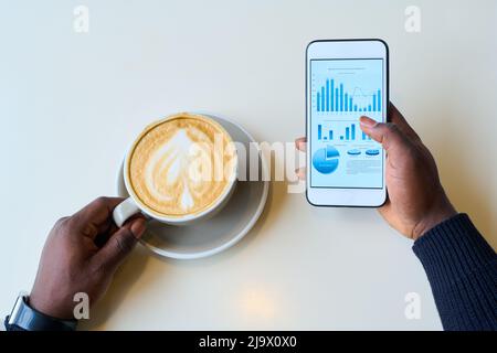 Blick aus der Perspektive eines afrikanischen Geschäftsmannes, der die Finanzmärkte auf dem Mobiltelefon untersucht, während er Cappuccino trinkt Stockfoto