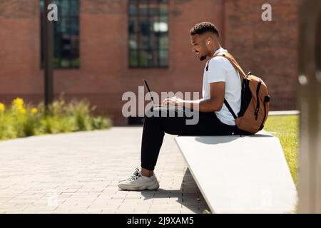 Schwarzer Mann in schnurlosen Ohrhörern, der im Freien einen Laptop verwendet Stockfoto