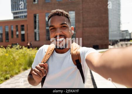 Glücklicher junger Mann, der Selfie-Bilder in Ohrhörern macht, die draußen spazieren gehen Stockfoto