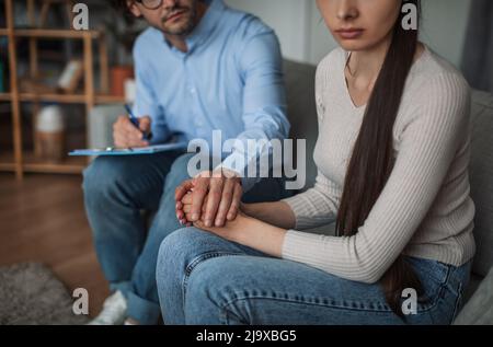 Junge europäische Kerl Arzt Psychologe mit Tablette hält Hand, beruhigt traurige Frau im Klinikinnenraum Stockfoto