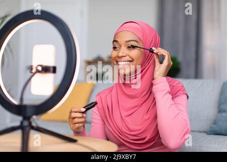 Online Make-up Lektion von Beauty-Blogger. Glückliche schwarze Dame, die Mascara auf die Wimpern aufsetzt und auf das Smartphone schaut Stockfoto