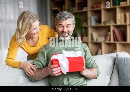 Liebevolle Frau überrascht ihren Mann mittleren Alters mit Geschenk zu Hause Stockfoto