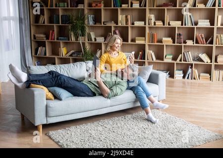 Glückliche Ehegatten Mittleren Alters Entspannen Mit Modernen Gadgets Auf Der Couch Zu Hause Stockfoto