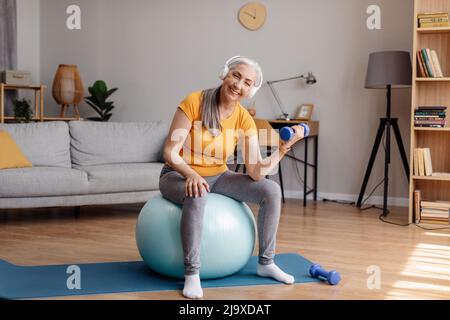 Aktive ältere Frau, die mit Hantel trainiert, auf einem Fitball sitzt, Musik in Kopfhörern hört, zu Hause trainiert Stockfoto