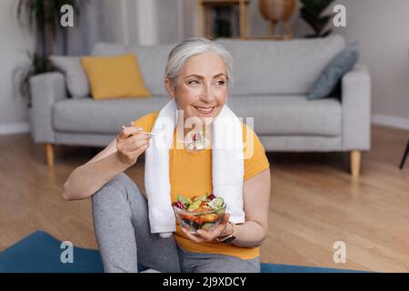 Gesundes Lifestyle-Konzept. Glückliche ältere Frau, die frischen Gemüsesalat isst und nach dem Training zu Hause auf einer Yogamatte sitzt Stockfoto