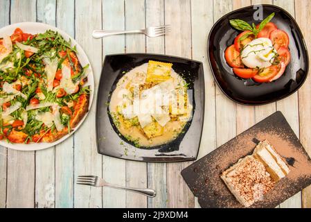 Italienische Gerichte mit Pasta, Burrata-Salat mit Tomaten und Olivenöl und neapolitanische Pizza mit Rucola, Parmesankäse, Kirschtomaten und Tirami Stockfoto