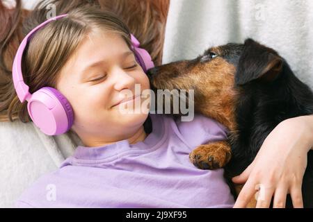 Fröhliches Teenager-Mädchen im lila T-Shirt in rosa Kopfhörern hört Musik und hat Spaß beim Spielen mit Hund Stockfoto