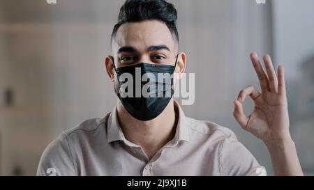 Glücklicher hispanischer männlicher Angestellter arabischer Mann Unternehmer tragen medizinische Maske verhindern Coronavirus Epidemie Blick auf Kamera zeigt okay Zeichen alles Stockfoto