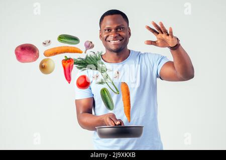 afroamerikanischer Koch kitchener hält eine Bratpfanne Zauberer Mann Kochen Magie fliegende Lebensmittel Salat, Karotte, Knoblauch, Zwiebel, Pfeffer, Kartoffel, Gurke Stockfoto