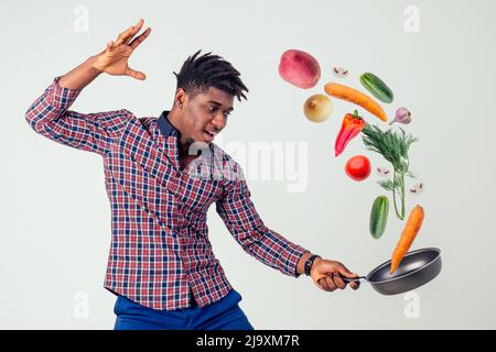 afroamerikanischer Koch kitchener hält eine Bratpfanne Zauberer Mann Kochen Magie fliegende Lebensmittel Salat, Karotte, Knoblauch, Zwiebel, Pfeffer, Kartoffel, Gurke Stockfoto