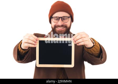 Bärtiger Kaukasier mit Brille und brauner Beanie lächelt und zeigt der Kamera auf weißem Hintergrund eine getönte Tafel. Hochwertige Fotos Stockfoto