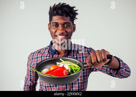 afroamerikanischer Koch kitchener hält eine Bratpfanne Zauberer Mann Kochen Magie fliegende Lebensmittel Salat, Karotte, Knoblauch, Zwiebel, Pfeffer, Kartoffel, Gurke Stockfoto