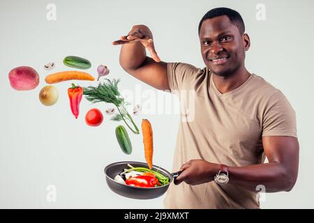 afroamerikanischer Koch kitchener hält eine Bratpfanne Zauberer Mann Kochen Magie fliegende Lebensmittel Salat, Karotte, Knoblauch, Zwiebel, Pfeffer, Kartoffel, Gurke Stockfoto