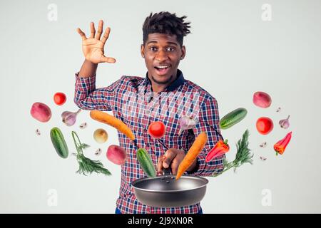 afroamerikanischer Koch kitchener hält eine Bratpfanne Zauberer Mann Kochen Magie fliegende Lebensmittel Salat, Karotte, Knoblauch, Zwiebel, Pfeffer, Kartoffel, Gurke Stockfoto