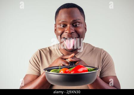 afroamerikanischer Koch kitchener hält eine Bratpfanne Zauberer Mann Kochen Magie fliegende Lebensmittel Salat, Karotte, Knoblauch, Zwiebel, Pfeffer, Kartoffel, Gurke Stockfoto