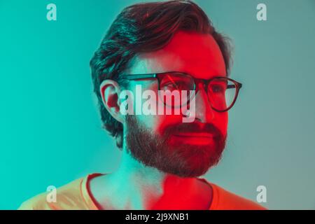 Nahaufnahme des Studios Porträt eines nachdenklichen, selbstbewussten, tausendjährigen europäischen bärtigen Mannes, der seine neue Brille mit rotem Neonlicht auf seinem Gesicht trägt. Brillenwerbe. Hochwertige Fotos Stockfoto
