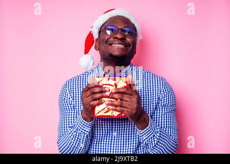 Afroamerikanischer Mann trägt stilvolles kariertes Hemd großes Lächeln in weihnachtsmütze mit Geschenkbox auf rosa Hintergrund Studio.dunkelhäutiger Weihnachtsmann Stockfoto