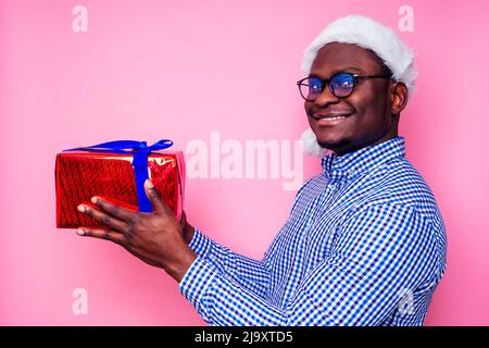 Afroamerikanischer Mann trägt stilvolles kariertes Hemd großes Lächeln in weihnachtsmütze mit Geschenkbox auf rosa Hintergrund Studio.dunkelhäutiger Weihnachtsmann Stockfoto