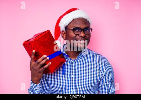 Afroamerikanischer Mann trägt stilvolles kariertes Hemd großes Lächeln in weihnachtsmütze mit Geschenkbox auf rosa Hintergrund Studio.dunkelhäutiger Weihnachtsmann Stockfoto