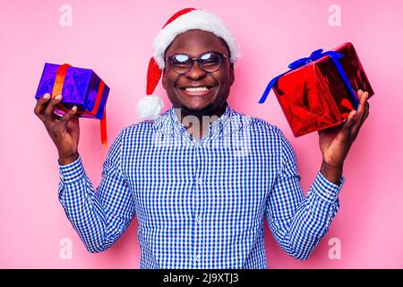 Afroamerikanischer Mann trägt stilvolles kariertes Hemd großes Lächeln in weihnachtsmütze mit Geschenkbox auf rosa Hintergrund Studio.dunkelhäutiger Weihnachtsmann Stockfoto