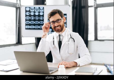 Porträt eines positiv selbstbewussten kaukasischen männlichen professionellen Arztes, der einen medizinischen Mantel mit Brille und Stethoskop trägt, in einem medizinischen Büro sitzt, die Kamera anschaut und freundlich lächelt Stockfoto