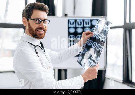 Gutaussehender, positiver kaukasischer Arzt, der Röntgenaufnahmen in seinem Schrank hält, ein Therapeut in medizinischer Uniform und eine Brille, die die Röntgenaufnahmen des Patienten lernt, in seiner Arztpraxis steht, die Kamera anschaut und lächelt Stockfoto