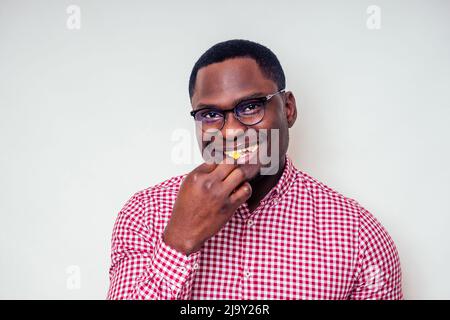 Schönes und junges afroamerikanisches männliches Modell in einem karierten rosa Hemd und einer stilvollen Brille, die Pommes frites frisst.Afro-Mann und Junk-Food auf Weiß Stockfoto