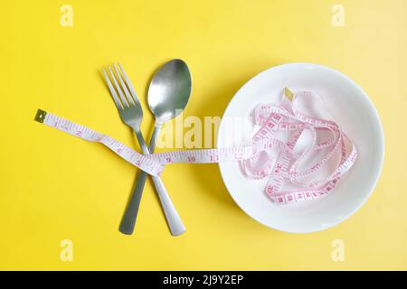 Gabel und Löffel mit Messband umwickelt flach in gelbem Hintergrund liegen. Anorexie, Ernährung und Ernährung, Gewicht zu verlieren Konzept. Stockfoto